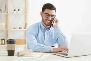 A smartly dressed man is sitting at his desk smiling while on the phone. He’s just starting his career in SaaS sales.